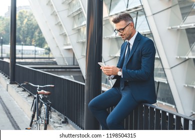 smiling middle aged businessman in eyeglasses using smartphone while sitting on street - Powered by Shutterstock