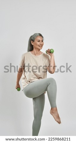 Smiling middle aged caucasian sportswoman raising legs and doing exercise with dumbbells. Modern healthy lifestyle. Female wearing sportswear. Isolated on white background. Studio shoot. Copy space