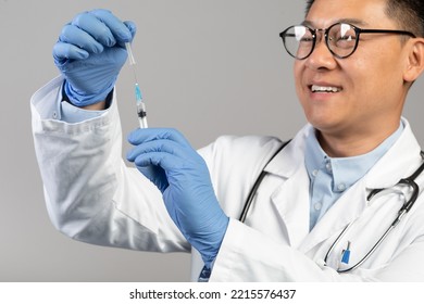 Smiling Middle Aged Chinese Male Doctor In White Coat And Glasses Take Vaccine Into Syringe, Isolated On Blue Background, Studio, Close Up. Immunization, Health Care, Treatment And Medicine, Covid-19