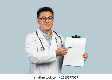 Smiling Middle Aged Chinese Male Doctor In White Coat And Glasses Shows On Tablet With Empty Space, Isolated On Blue Background, Studio. Prescription For Treatment, Health Care, Medicine, Offer And Ad