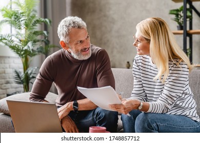 Smiling Middle Aged Couple With Laptop Studying Documents While Working On Couch At Home