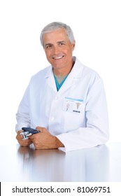 Smiling Middle Aged Doctor Seated And Holding His Stethoscope In Both Hands. Man Is Wearing A Lab Coat And Scrubs In Vertical Format Over A White Background.