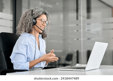 Smiling middle aged female support service employee talking to customer sitting at desk. Happy senior old woman professional call contact center agent wearing headset hybrid working in business office - Powered by Shutterstock