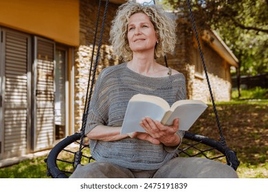 Smiling middle aged good-looking blond curly haired woman, reading a book in hammock in the garden during spring, summer. - Powered by Shutterstock