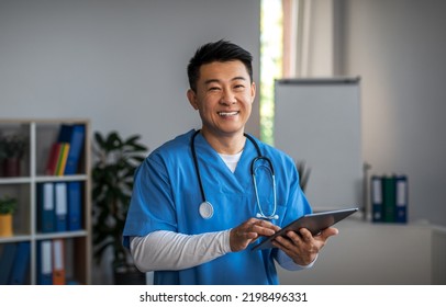 Smiling Middle Aged Japanese Male Doctor With Tablet And Wait Patient In Clinic Office Interior. Treatment With Professional Therapist, Medical Health Care And Good Service During Covid-19 Pandemic