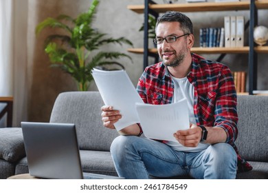 Smiling middle aged man in glasses reading paper documents, letters, calculating bills while working laptop and using online bank service at home. Freelancer looking at contract with good offer - Powered by Shutterstock