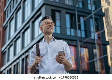 Smiling middle aged man using smartphone, standing on the street. Mobile banking concept. Portrait of handsome mature businessman holding messenger bag, wearing stylish eyeglasses looking at camera - Powered by Shutterstock