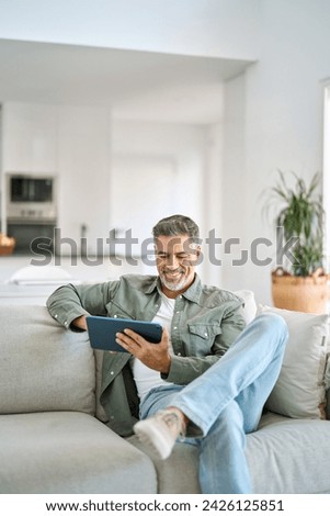 Smiling middle aged man using digital tablet relaxing on couch at home. Happy mature male user holding tab computer browsing internet on pad technology device sitting on sofa in living room. Vertical.