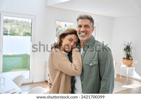 Smiling middle aged mature couple embracing standing at home. Happy romantic affectionate senior older man and woman in love enjoying hugging looking at camera in modern house living room. Portrait.