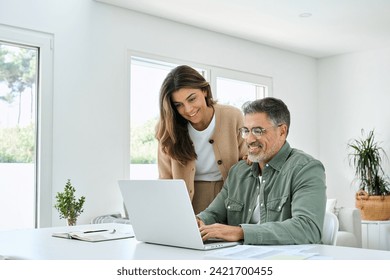 Smiling middle aged senior man working on computer sitting at table with wife standing nearby in living room. Happy mature older couple using laptop technology at home. Authentic candid shot. - Powered by Shutterstock