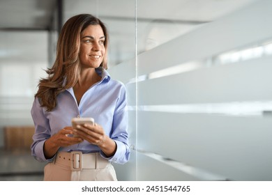 Smiling middle aged woman business investor using mobile banking apps on cellular phone standing in office. Mature businesswoman executive holding cellphone working on smartphone looking away. - Powered by Shutterstock