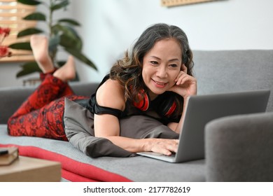Smiling middle aged woman chatting online in social network, watching movie on laptop while lying on couch