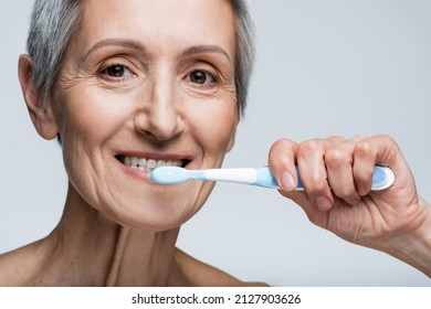 Smiling Middle Aged Woman Holding Toothbrush And Brushing Teeth Isolated On Grey