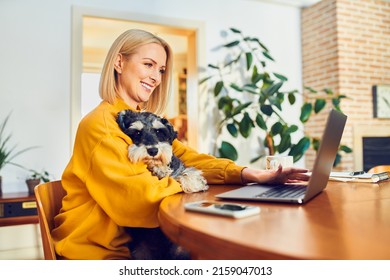 Smiling Middle Aged Woman Holding Dog Using Laptop While At Home Office