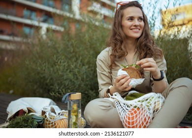 Smiling Middle Aged Woman In Overall With Tote Bag, String Bag And Sandwich Eating While Sitting Outside In The City.