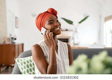 Smiling Middle Aged Woman Sitting On Couch At Home Talking Over Smartphone. Happy Black Woman With Traditional Head Turban Using Mobile Phone For Conversation. Relaxing Mid African American Lady.