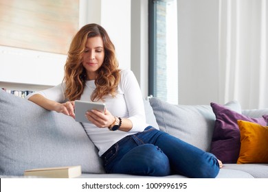 A Smiling Middle Aged Woman Using Digital Tablet While Relaxing On Sofa At Home.