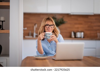 Smiling middle aged woman using laptop and having video call while working from home. Confident female drinking tea while sitting at table at the kitchen.