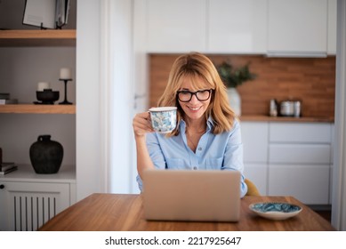 Smiling middle aged woman using laptop and having video call while working from home. Confident female drinking tea while sitting at table at the kitchen.