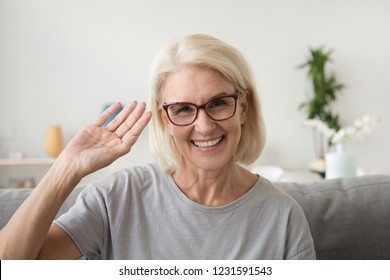 Smiling middle aged woman waving hand looking at camera, older mature lady in glasses making video blog or call at home, happy friendly senior vlogger sitting on sofa dating online, headshot portrait