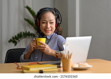 Smiling Middle Aged Woman Wearing Headphone Watching Online Webinar On Laptop Computer At Home Kitchen