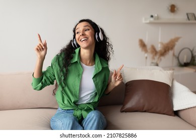Smiling Middle Eastern Lady Wearing Headphones Listening To Music And Favorite Songs Relaxing Sitting On Couch At Home. Weekend Leisure And Relaxation. Great Playlist Concept - Shutterstock ID 2107825886