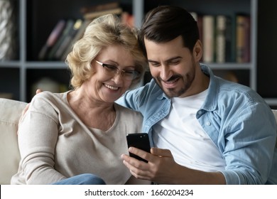Smiling middle-aged 60s mother rest with grown-up son using smartphone together, happy young man enjoy family weekend with senior mom browsing wireless Internet on cellphone, have fun at home