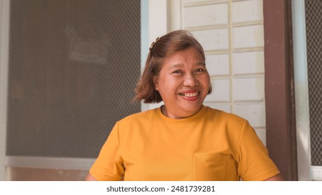 Smiling middle-aged asian woman in a yellow shirt standing outside her townhouse. Bright, cheerful expression - Powered by Shutterstock