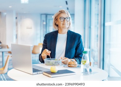 Smiling middle-aged business woman having healthy lunch at working place or business cafe, with laptop during her break. Balanced diet lunch box. Healthy eating habits and well-being. Selective focus - Powered by Shutterstock