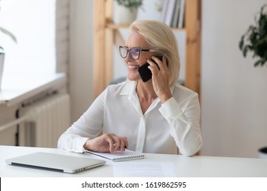 Smiling middle-aged businesswoman sit at desk smiles talking on smartphone looking out the window enjoy pleasant conversation with client, female sales manager makes successful profitable deal concept - Powered by Shutterstock