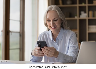 Smiling middle-aged businesswoman using smartphone seated at workplace, distracted from work chatting on internet, read message from client, check calendar. Modern tech for business, agenda concept