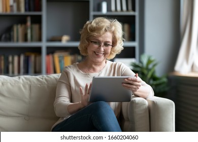  Smiling middle-aged Caucasian woman sit on couch in living room browsing wireless Internet on tablet, happy modern senior female relax on sofa at home using pad device, elderly technology concept