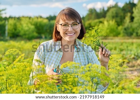 Smiling middle-aged female with pruner picking branches of flowering dill in garden.