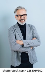 Smiling Middle-aged Grey-haired Man In Black Glasses, Standing With Arms Crossed, Wearing Black Sweater And Grey Blazer, Against Plain Blue Background With Shadow