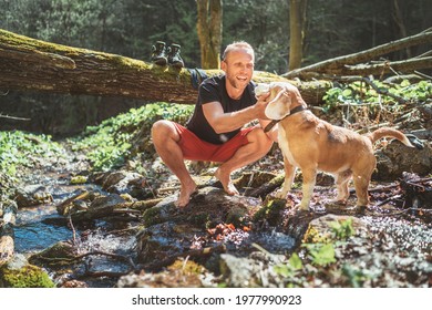 Smiling middle-aged man petting his beagle dog near mountain forest stream while he waiting for laundry drying and trekking boots. Active people traveling with pets concept image.