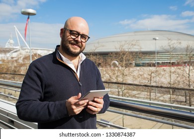 Smiling middle-aged man using tablet outdoors. Bold man wearing casual clothes, looking at camera and standing outdoors with stadium view in background. Lifestyle concept. Front view. - Powered by Shutterstock - Get 15% off with code: PIKWIZARD15