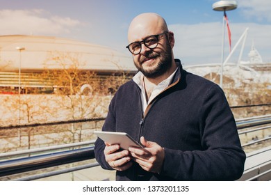 Smiling middle-aged man using tablet outdoors. Bold man wearing casual clothes, looking at camera and standing outdoors with stadium view in background. Lifestyle concept. Front view. - Powered by Shutterstock - Get 15% off with code: PIKWIZARD15