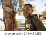 A smiling middle-aged Papuan sits against the backdrop of a Papuan village and the ocean