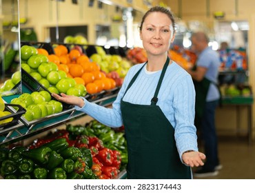 Smiling middle-aged shop assistant posing in front of counter in greengrocery with big stock