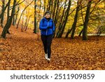 Smiling middle-aged woman dressed sporty in black leggings, blue jacket, black cap and sports backpack on her back running through forest full of fallen golden leaves on beautiful autumn day.