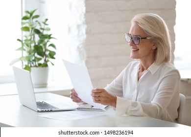 Smiling Middle-aged Woman In Glasses Sit At Office Desk Work At Laptop Reading Paper Document, Happy Senior Businesswoman In Spectacles Look Through Consider Paperwork Contract Receive Good News