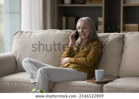 Smiling middle-aged woman holds cellphone makes call to grown up children or friend sit on sofa at home. Distance communication, older generation using modern wireless tech, phonecall activity concept