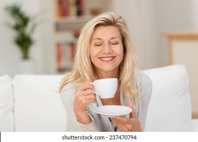 Smiling Middle-aged Woman Savouring Her Coffee Sitting Holding A Cup And Saucer On Her Sofa With Her Eyes Closed And Smile Of Bliss