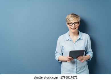 Smiling middle-aged woman with short blond hair, wearing glasses and blue shirt, standing with tablet computer in her hands and looking at camera. Front portrait on blue background with copy space