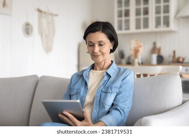 Smiling middle-aged woman sit on couch with digital tablet, browsing wireless internet, relaxing on sofa at home using pad device, doing freelance work, chatting remotely, check balance via e-bank app - Powered by Shutterstock