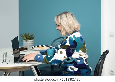 A smiling middle-aged woman working on a laptop at a dining table in a casual setting, wearing a bright turquoise sweater, with copy space.
 - Powered by Shutterstock