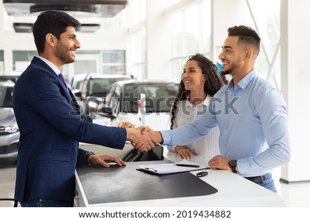 Smiling middle-eastern young man and woman buying car at auto salon, handsome arab guy shaking sales assistant hand, making successful deal, happy family got brand new luxury car, side view
