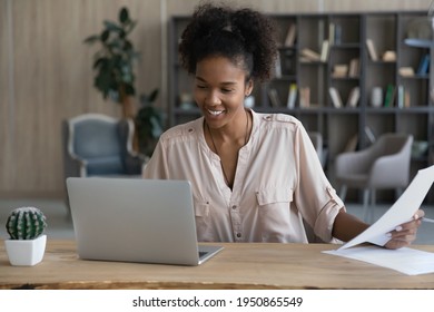 Smiling millennial 20s African American woman sit at desk at home office work online on computer with paper documents. Happy young biracial female study prepare research distant on laptop gadget.