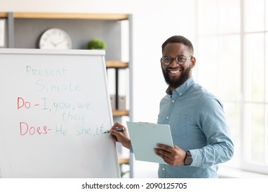 Smiling millennial african american bearded guy teacher in glasses stands near blackboard and teaches english in home interior. Remote tutoring, social distance study and knowledge during covid-19