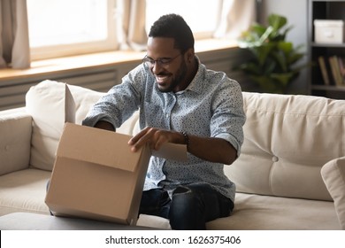 Smiling millennial african american man customer opening cardboard box sit on sofa at home, happy ethnic male consumer unpack parcel receive retail purchase fast postal shipping delivery concept